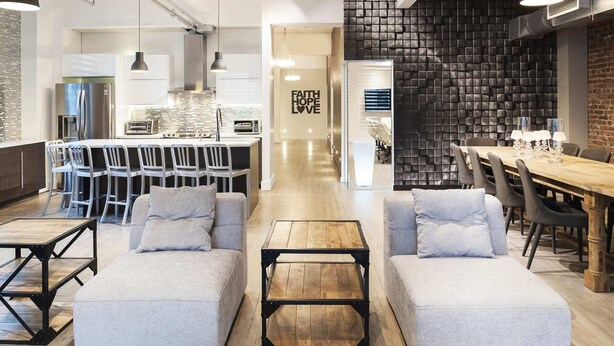 Un salon moderne avec un canapé, un pouf et une table basse, un coin repas avec des chaises et une table, et une cuisine en arrière-plan.