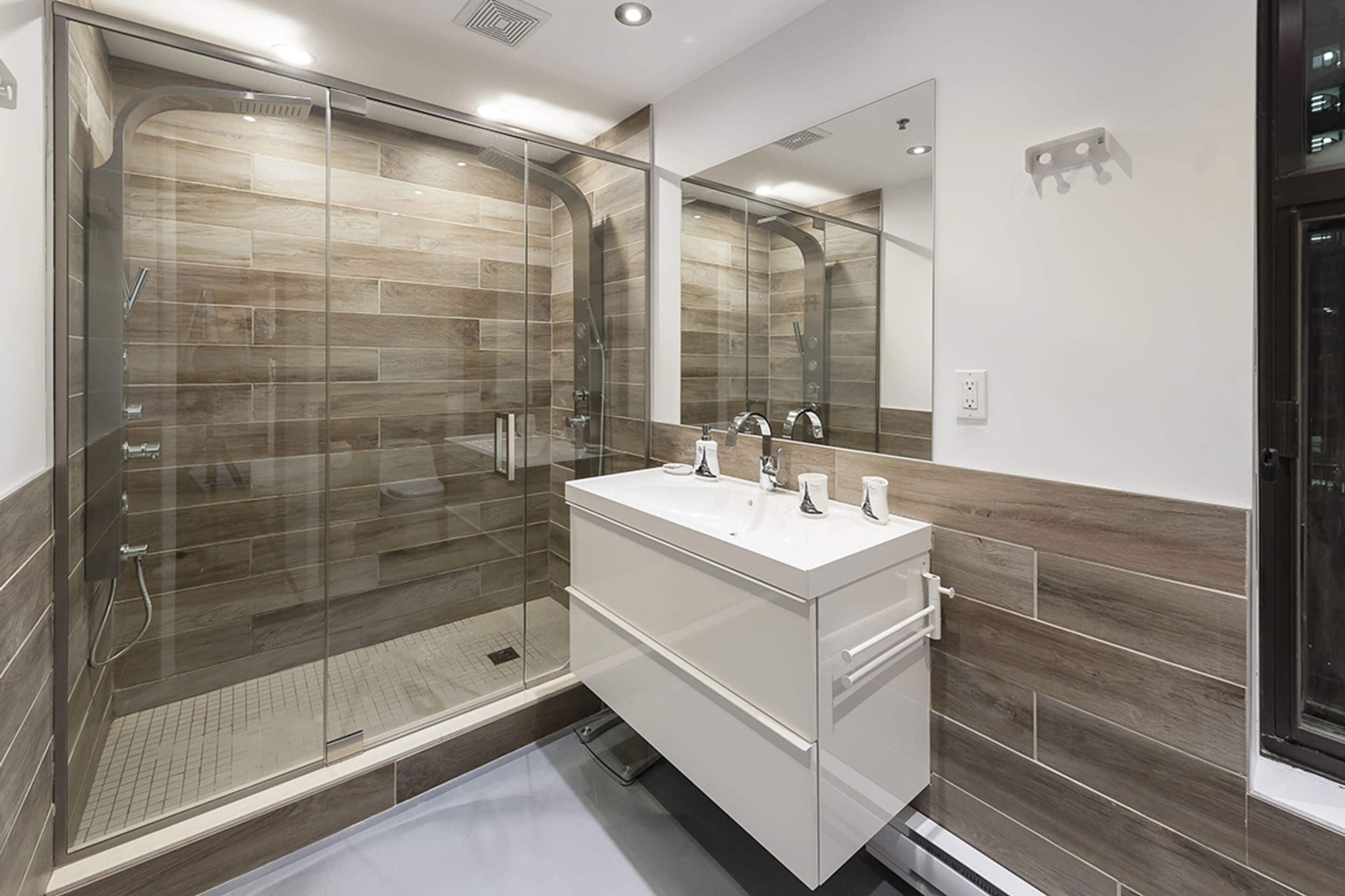 A modern bathroom with a glass-enclosed shower, a white vanity with a mirror, and wooden wall tiles.