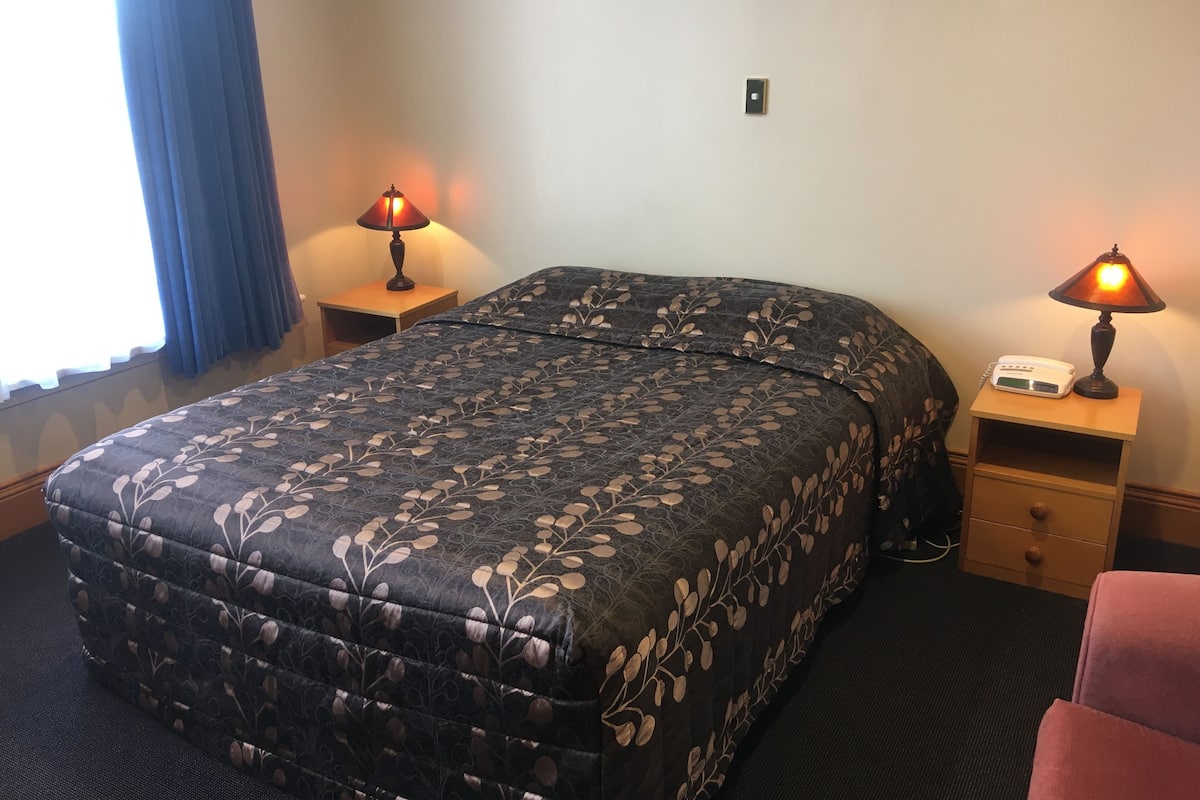 A hotel room with a large bed, two bedside tables with lamps, a small drawer, and a window with curtains.