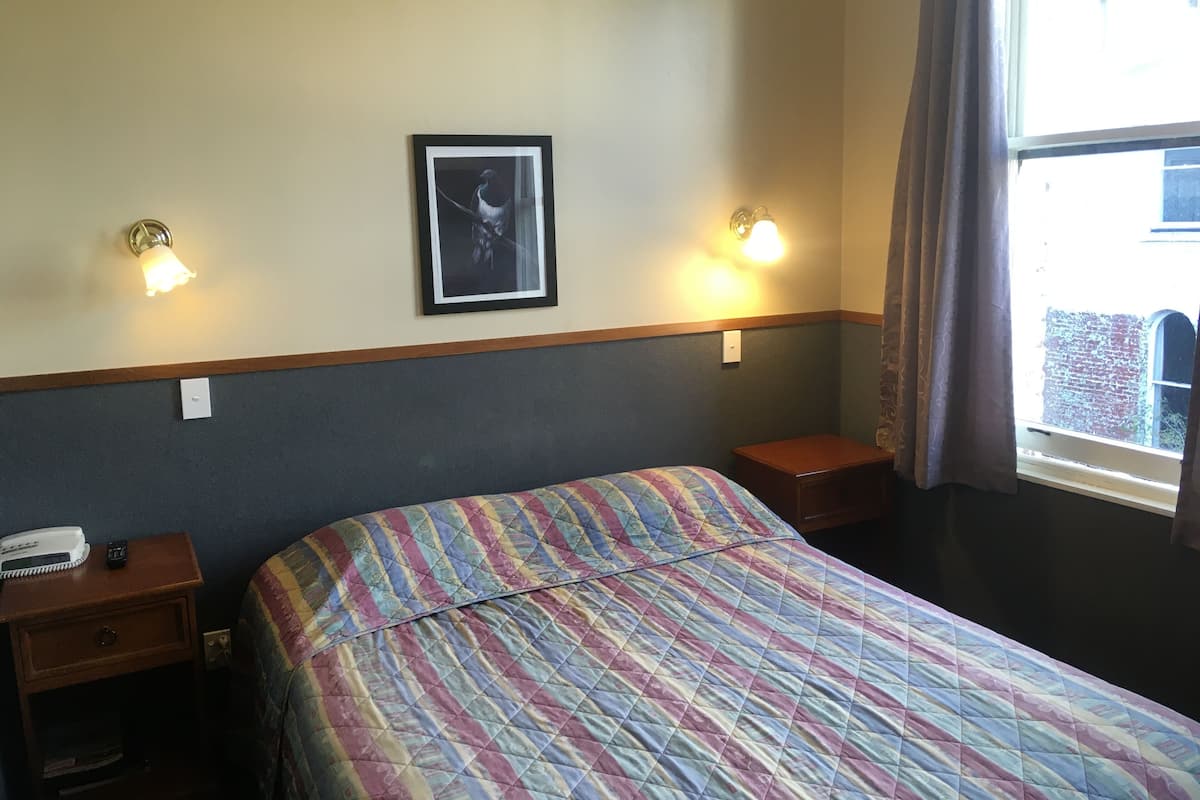 A hotel room with a bed, two bedside tables, a nightstand, a framed picture on the wall, and a window with curtains.