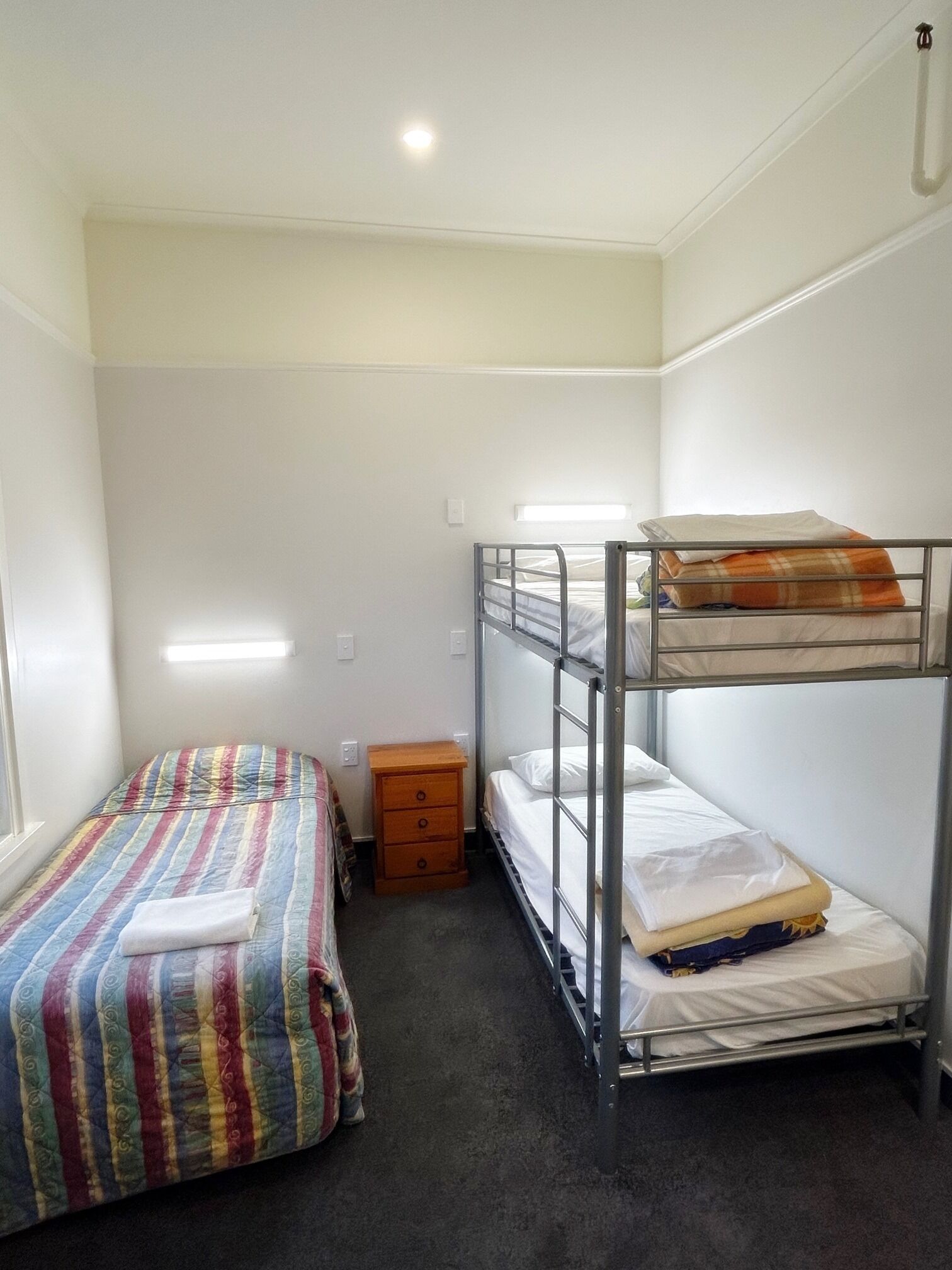 A dormitory room with bunk beds, a single bed, a small wooden nightstand, and a window.