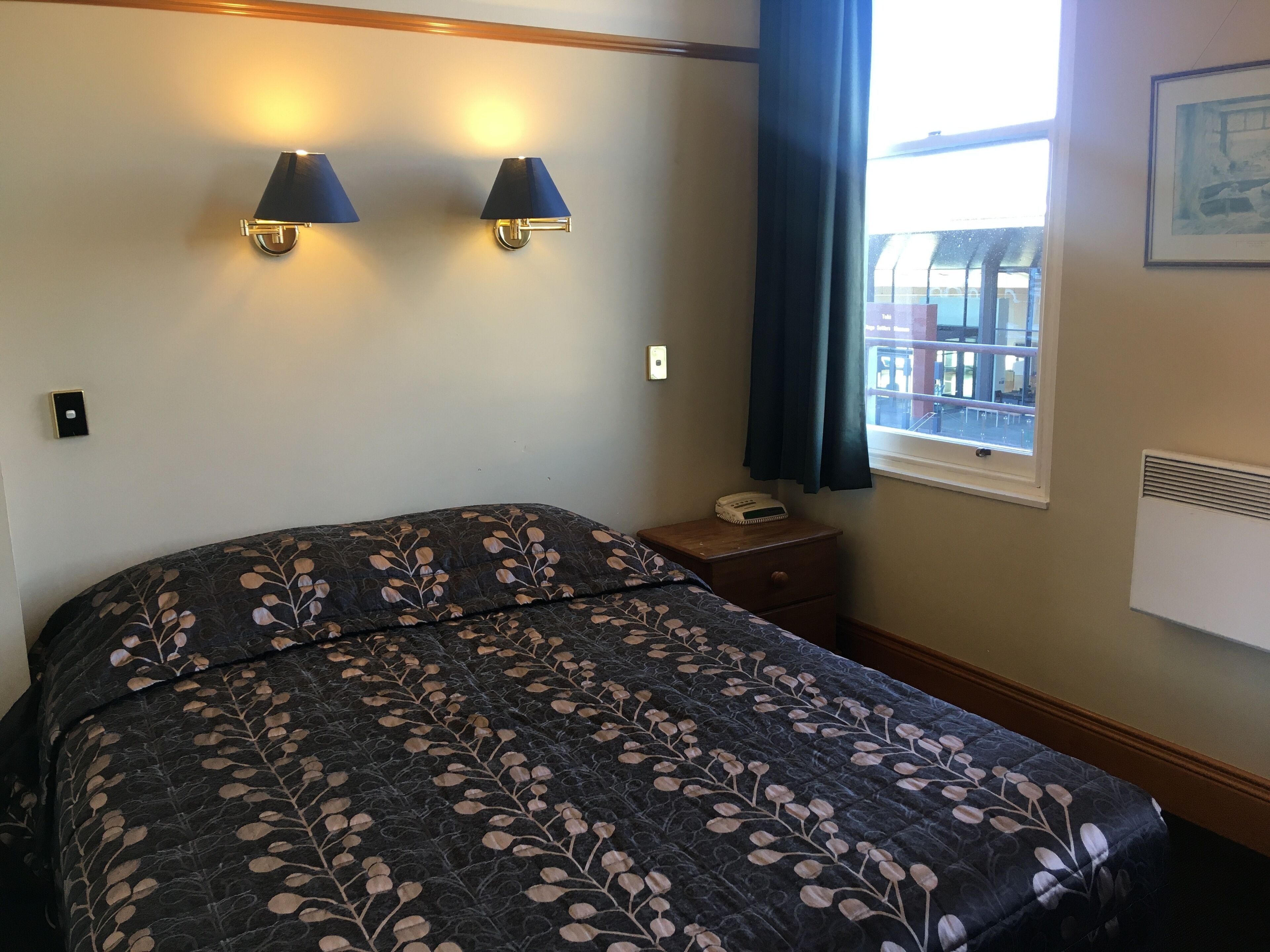 A hotel room with a large bed, two wall-mounted lamps, a bedside table with a phone, and a window with a view of buildings.