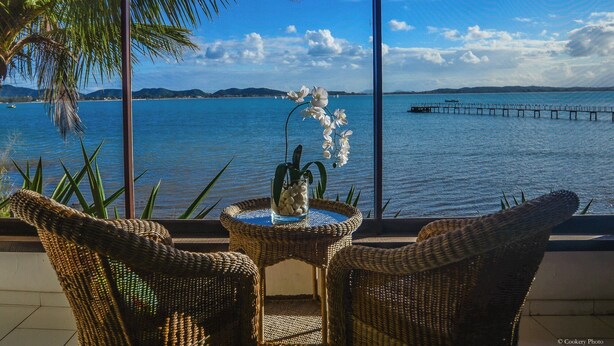 A balcony with wicker chairs and a table, overlooking a calm sea with a pier and distant hills. - Barracuda Resort (Búzios)