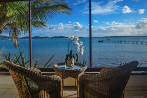 A balcony with wicker chairs and a table, overlooking a calm sea with a pier and distant hills. - Barracuda Resort (Búzios)
