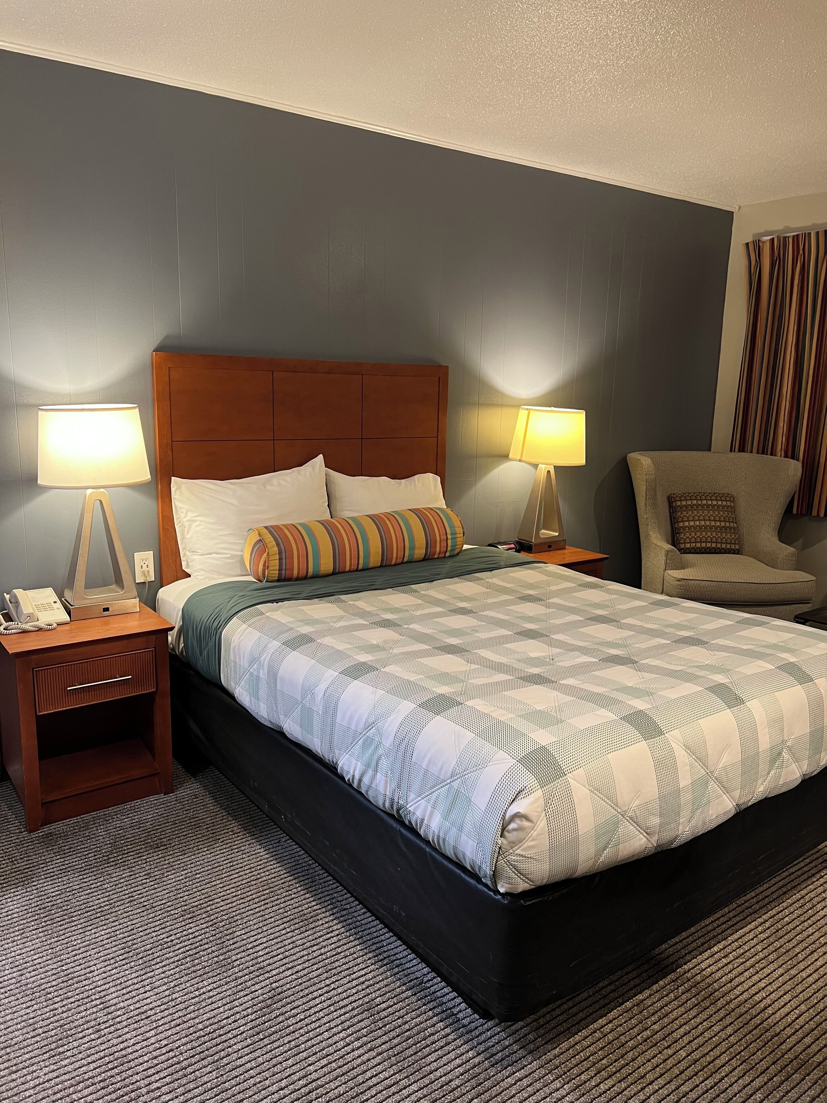 Habitación de hotel con una cama grande, dos mesitas de noche con lámparas, una silla y un teléfono a la izquierda.