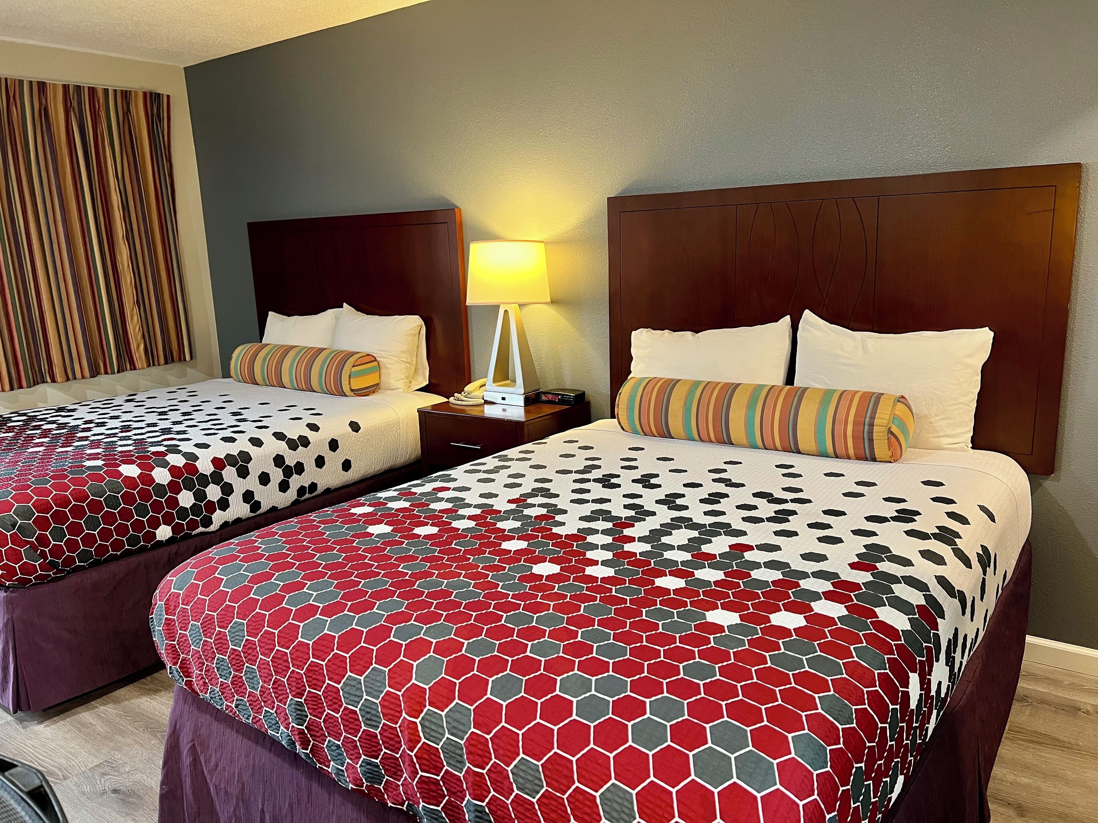 Two double beds with patterned bedding, a nightstand with a lamp, and a striped curtain.