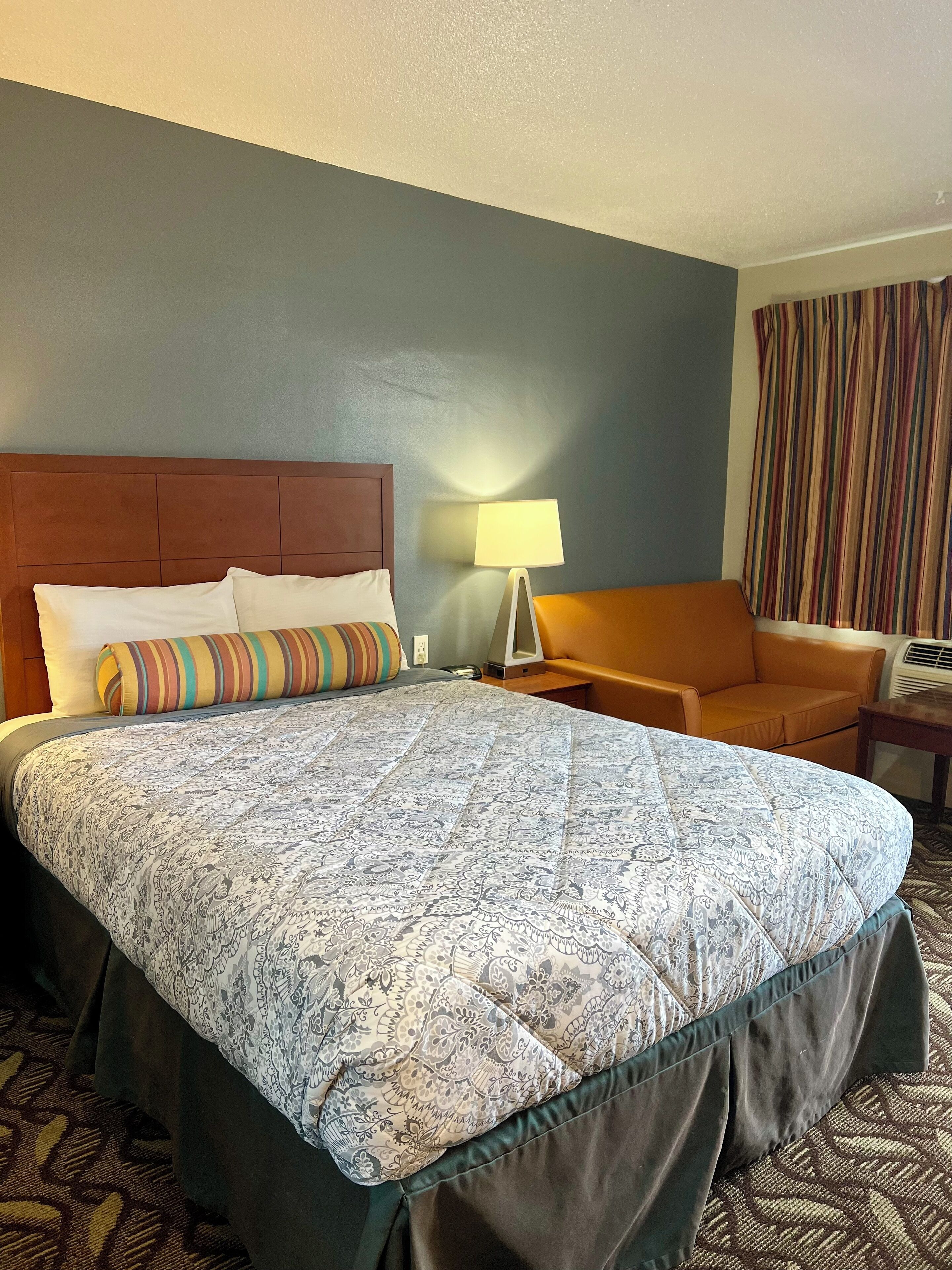 Una habitación de hotel con una cama grande, un cabecero de madera, un almohadón a rayas, una lámpara, un sillón naranja y una cortina a rayas.