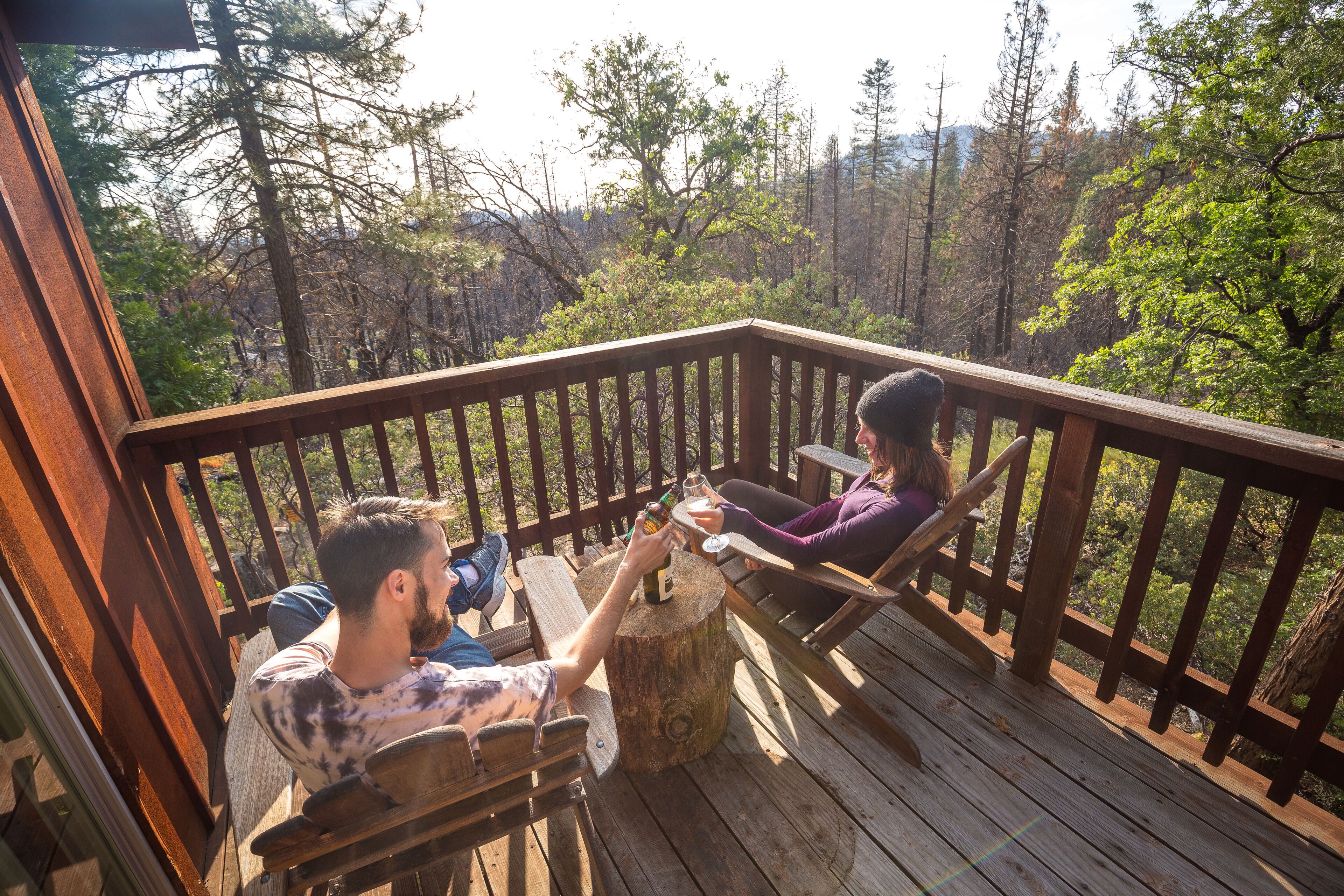 Eine Holzterrasse mit zwei Personen, die an einem Tisch sitzen, umgeben von einer bewaldeten Landschaft.