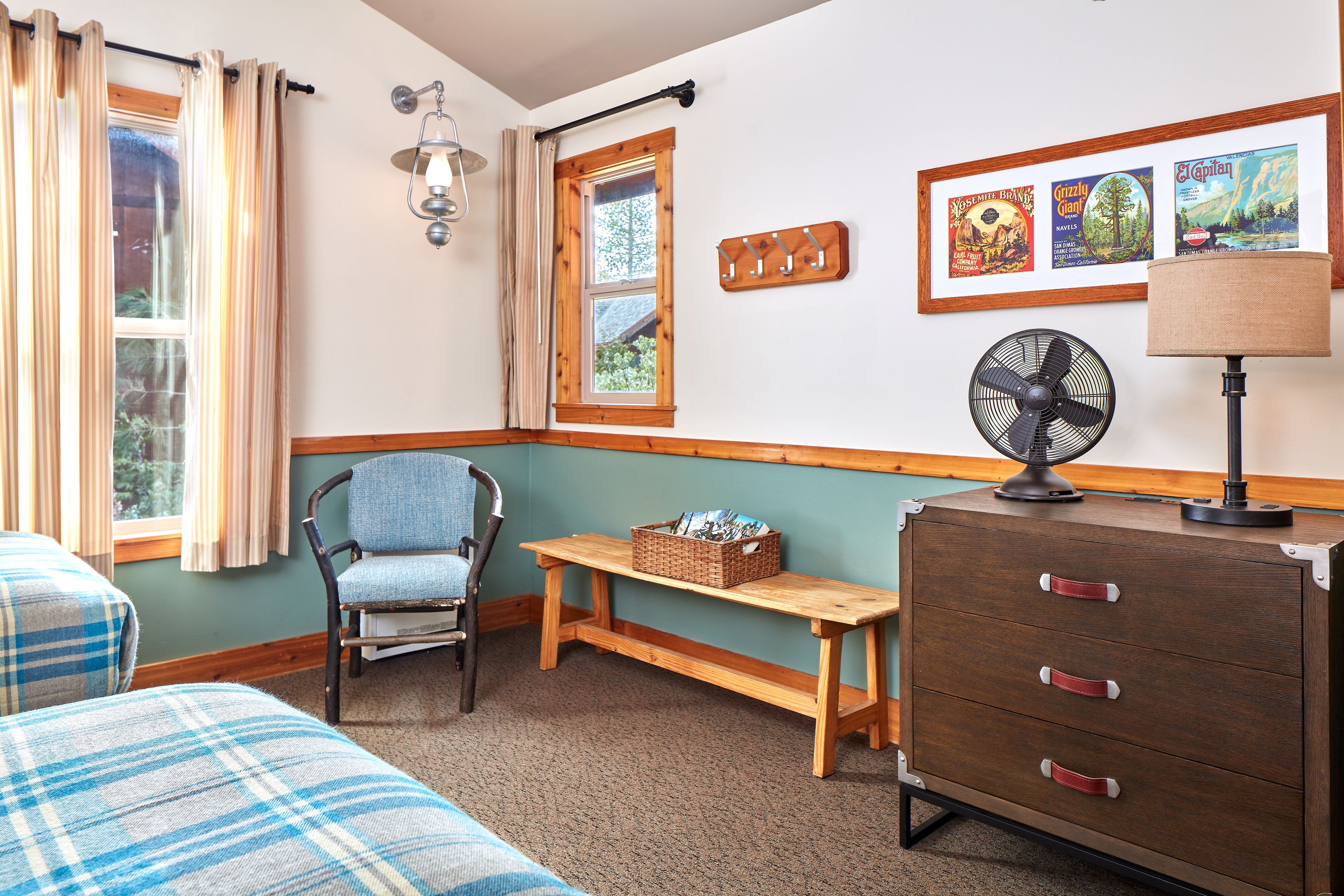 Une pièce avec deux lits, une chaise, un banc en bois, une commode, un ventilateur et des tableaux encadrés accrochés au mur.