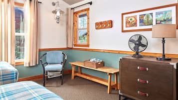 Une pièce avec deux lits, une chaise, un banc en bois, une commode, un ventilateur et des tableaux encadrés accrochés au mur.