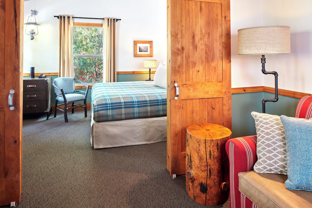 A hotel room with a bed, a desk, a chair, a lamp, and a wooden door.