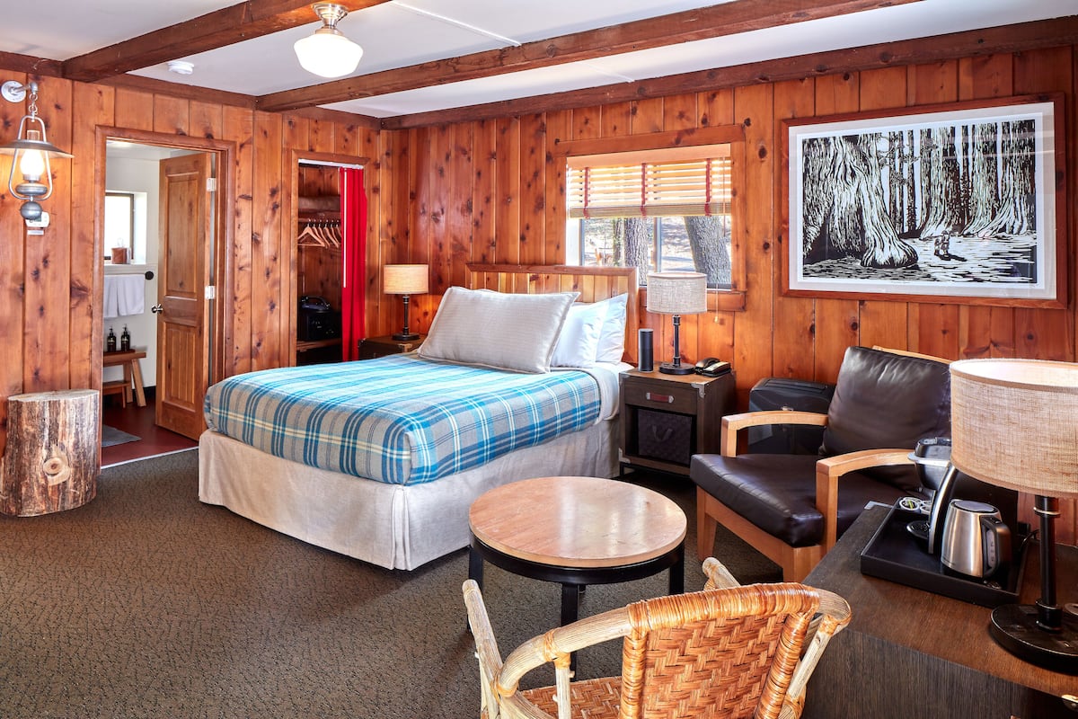 A wooden room with a bed, a chair, a table, and a painting on the wall.