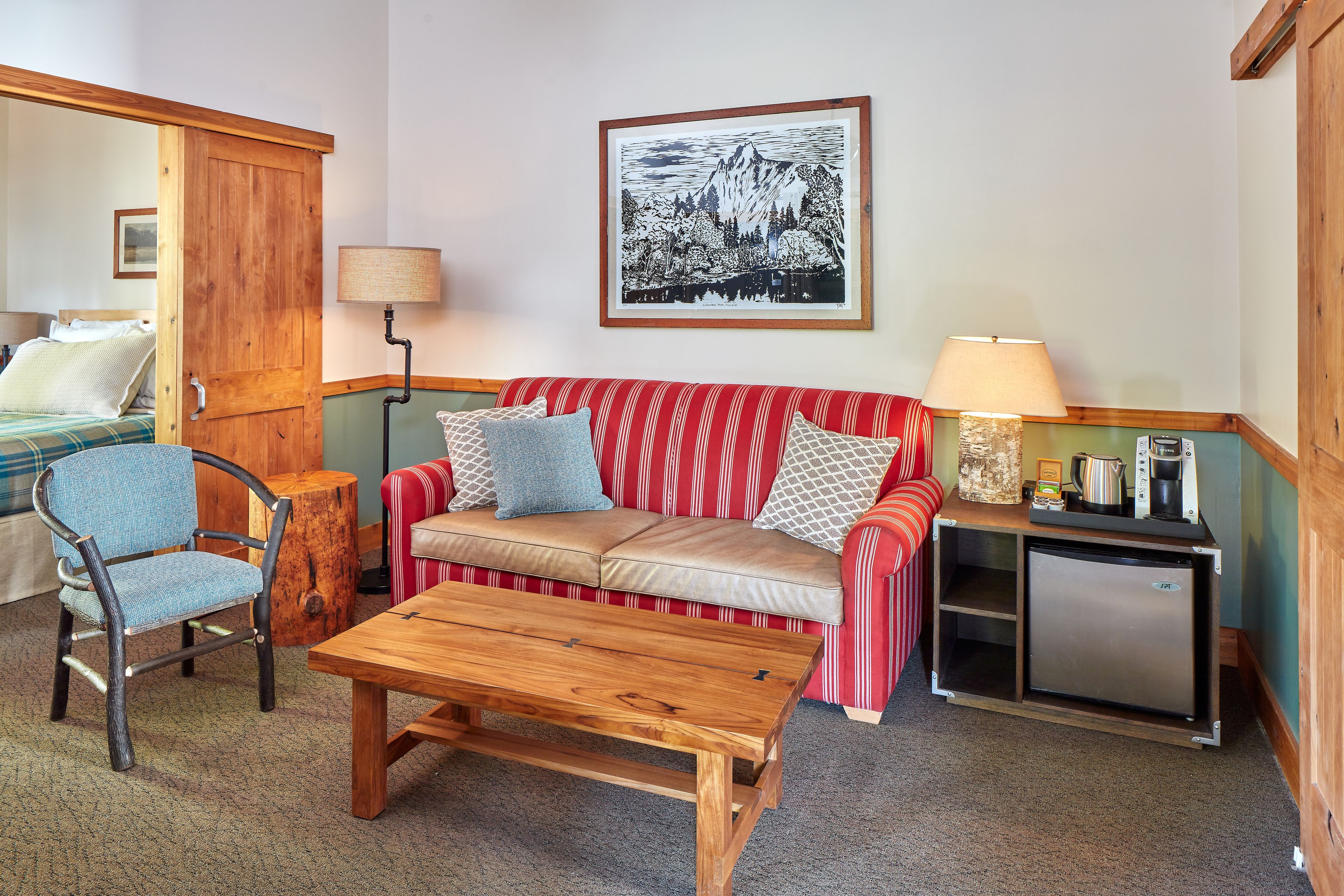 A cozy room with a wooden chair, a red-striped sofa, a small wooden table, a framed black and white landscape painting, and a compact kitchenette.