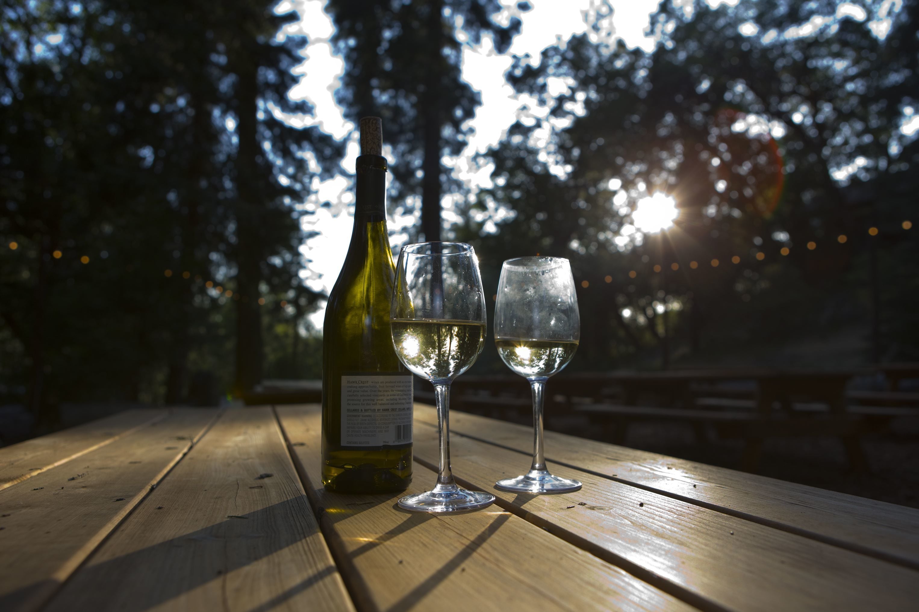 A wooden table with a bottle and two glasses of white wine, set outdoors with trees and sunlight in the background.