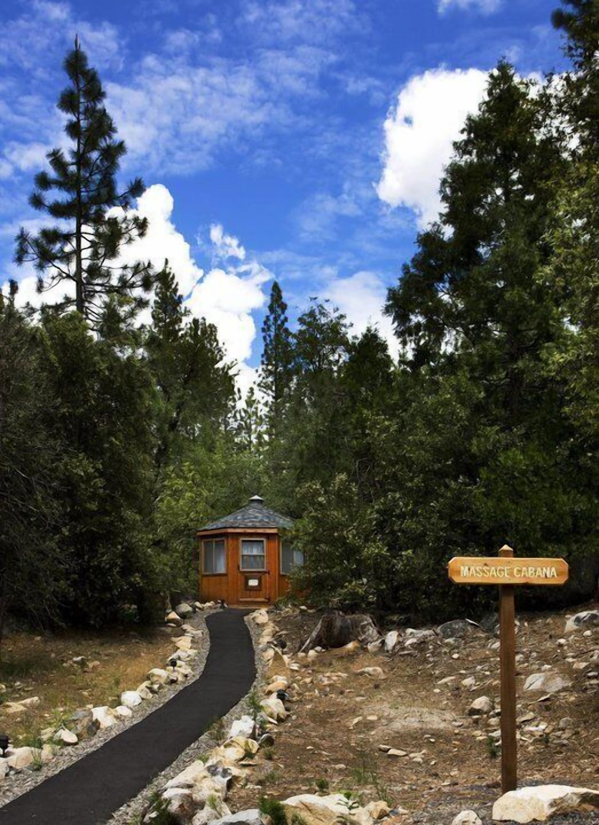 Una cabaña de madera con el rótulo 'Massage Cabana', rodeada de árboles y un sendero de piedra.
