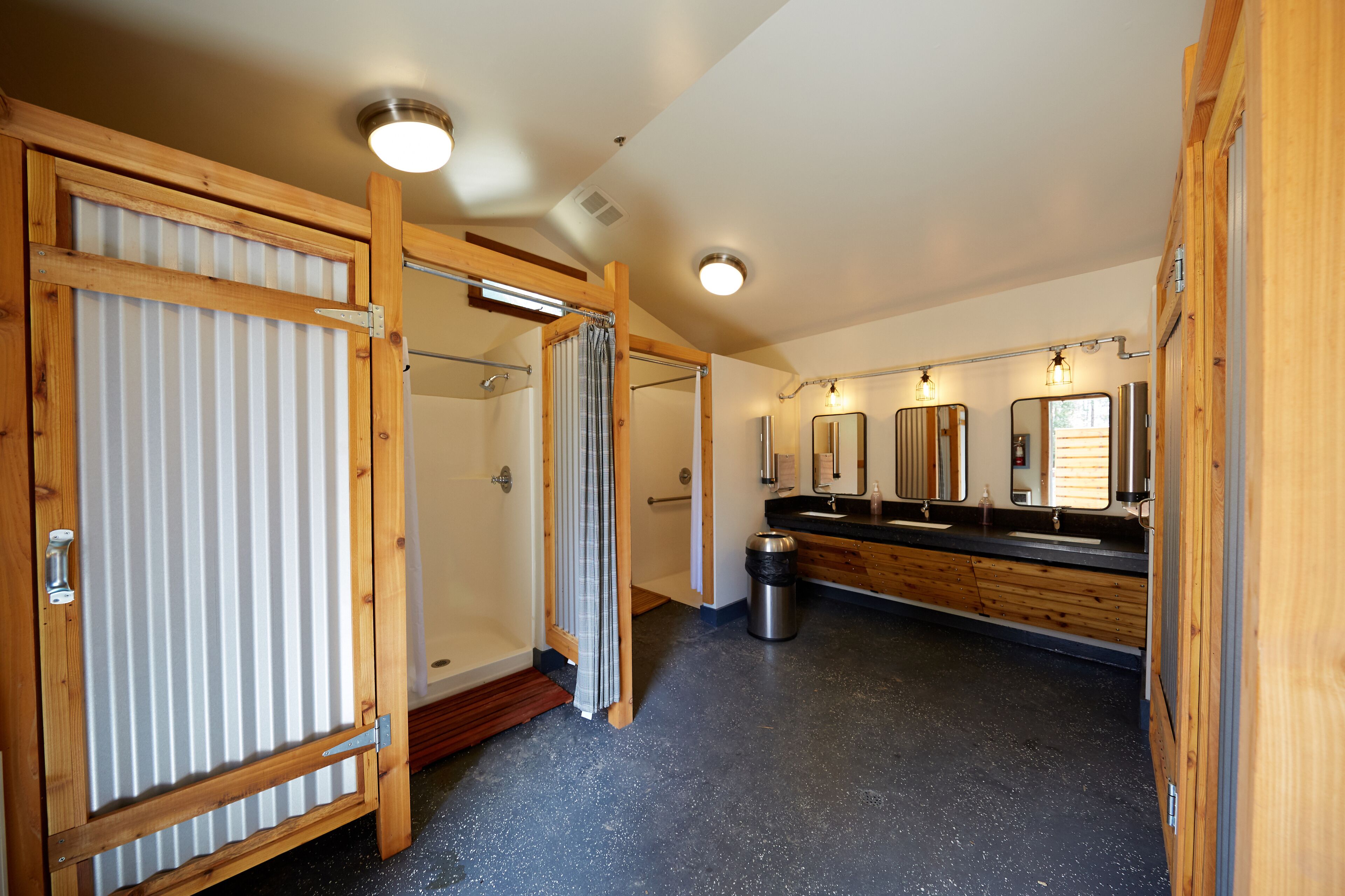 A bathroom with wooden doors, a shower area, a sink with a mirror, and a trash bin.