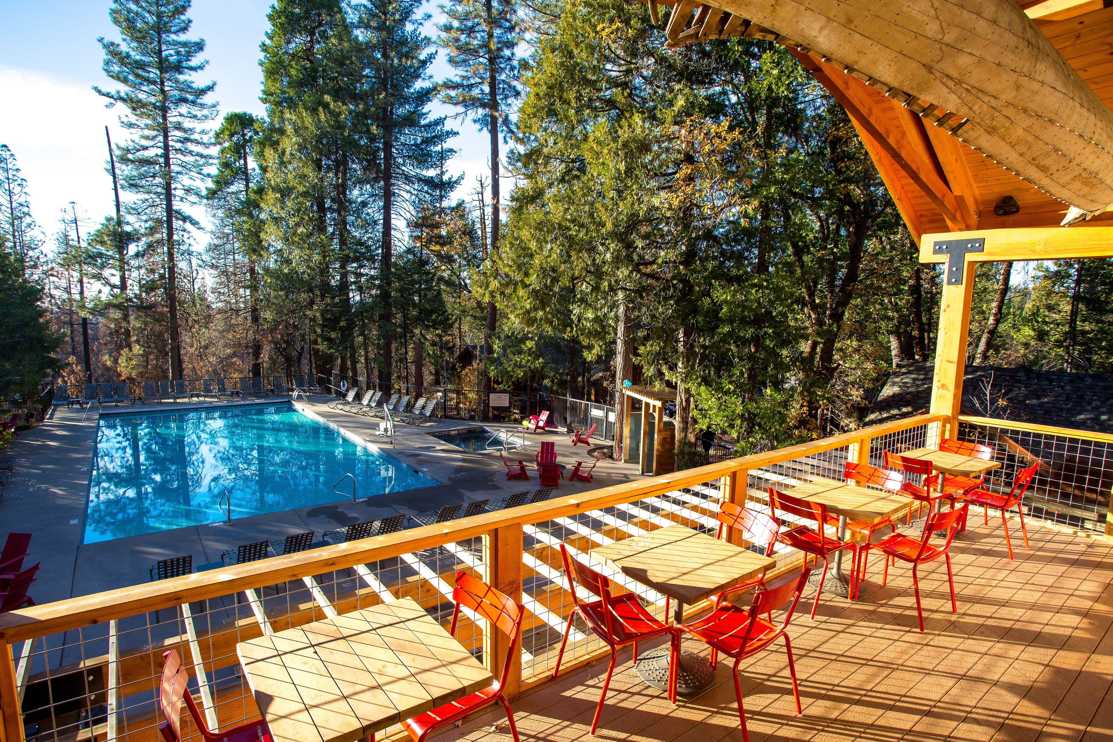 A pool area with a wooden deck, red chairs, and tables, surrounded by tall trees.