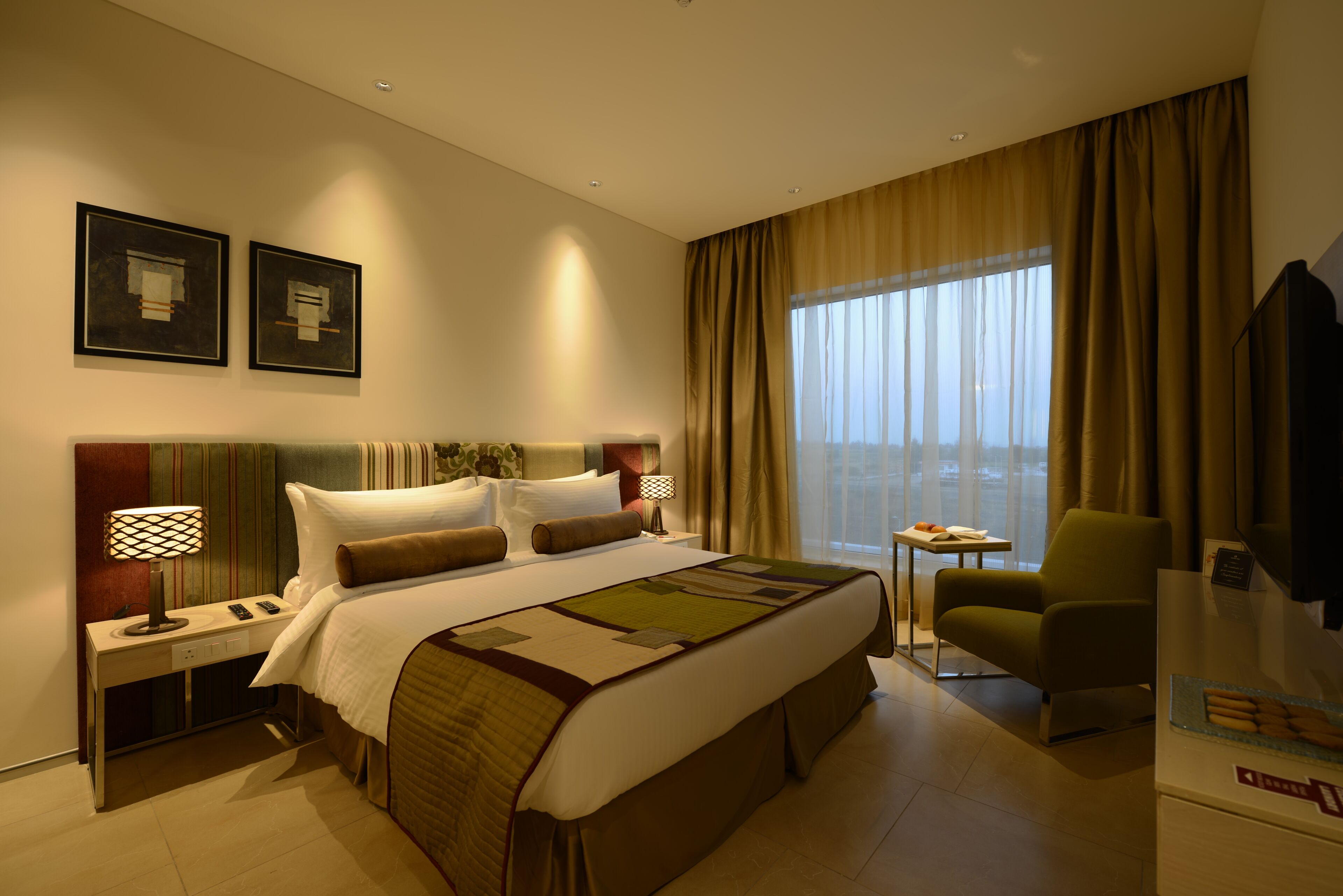 A hotel room with a large bed, a desk with a lamp, a chair, and a TV.