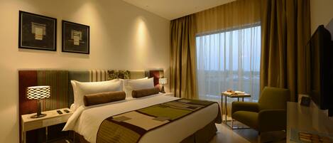 A hotel room with a large bed, a desk with a lamp, a chair, and a TV.