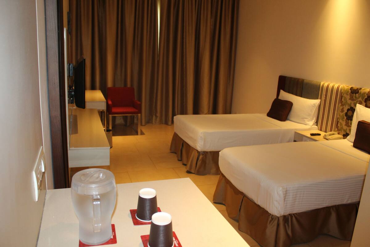 A hotel room with two single beds, a desk with a chair, a TV, and a large jug on the table.