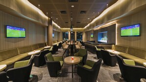 A modern lounge area with sofas, chairs, and tables, featuring large screens displaying sports events.