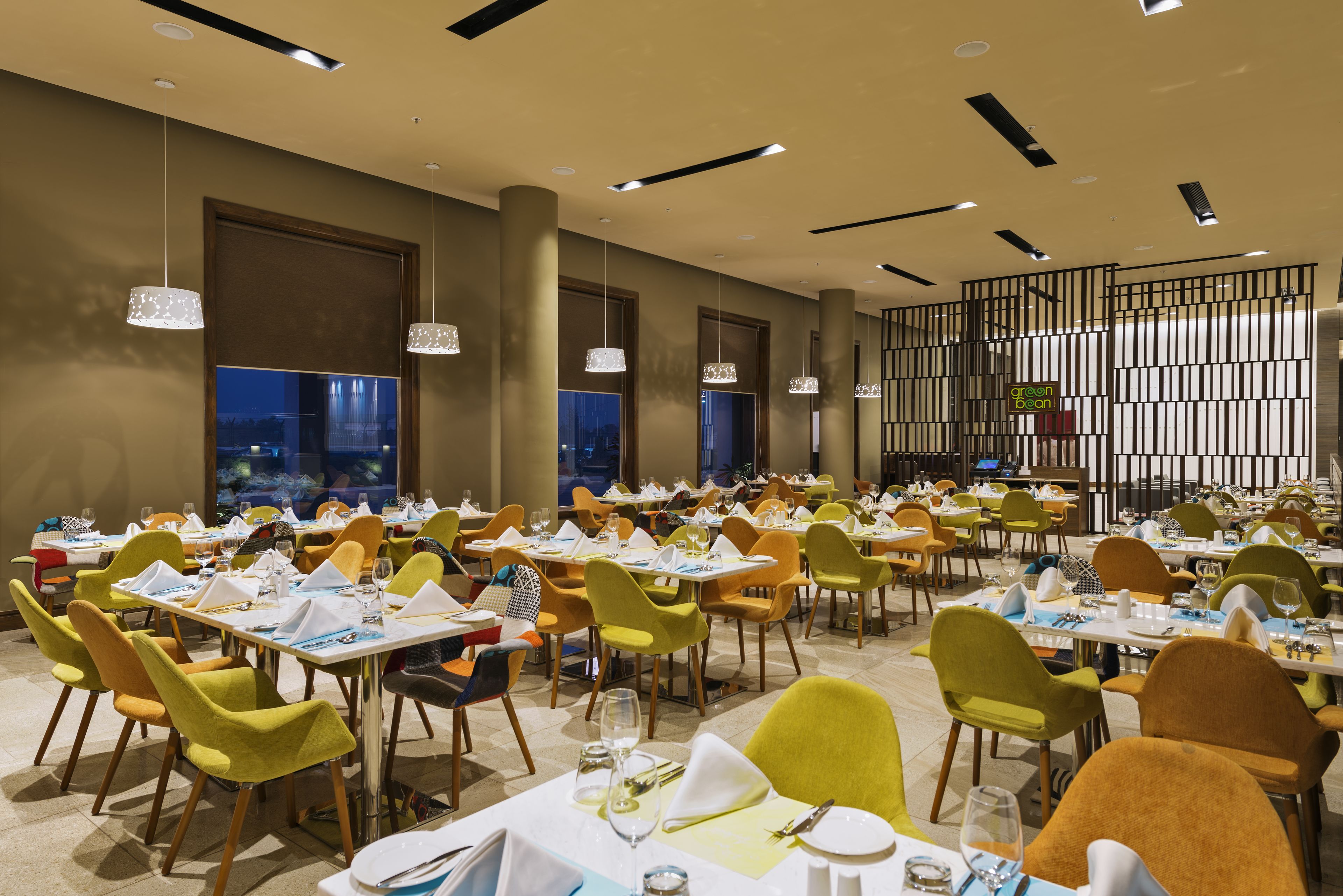 A modern dining area with multiple tables and chairs, featuring a mix of yellow and orange chairs, and pendant lighting.