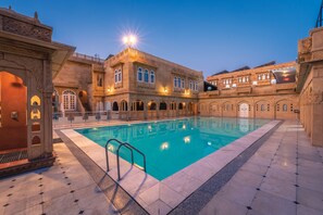 A pool area with a clear blue pool, surrounded by ornate buildings with arched windows and a tiled floor.