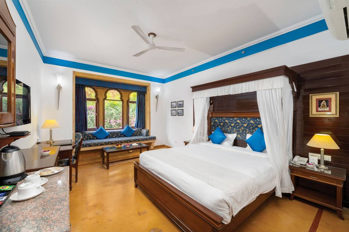 Una habitación de hotel con una cama grande, una zona de estar con un sofá y sillones, una mesa de comedor con tetera y tazas, y una ventana con cortinas.