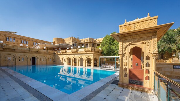 A pool area with a clear blue pool, surrounded by ornate architecture and a tiled floor. - Rang Mahal Jaisalmer (Jaisalmer)