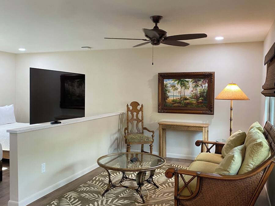 A living room with a glass-top coffee table, a wicker sofa, a television, a ceiling fan, and a painting on the wall.