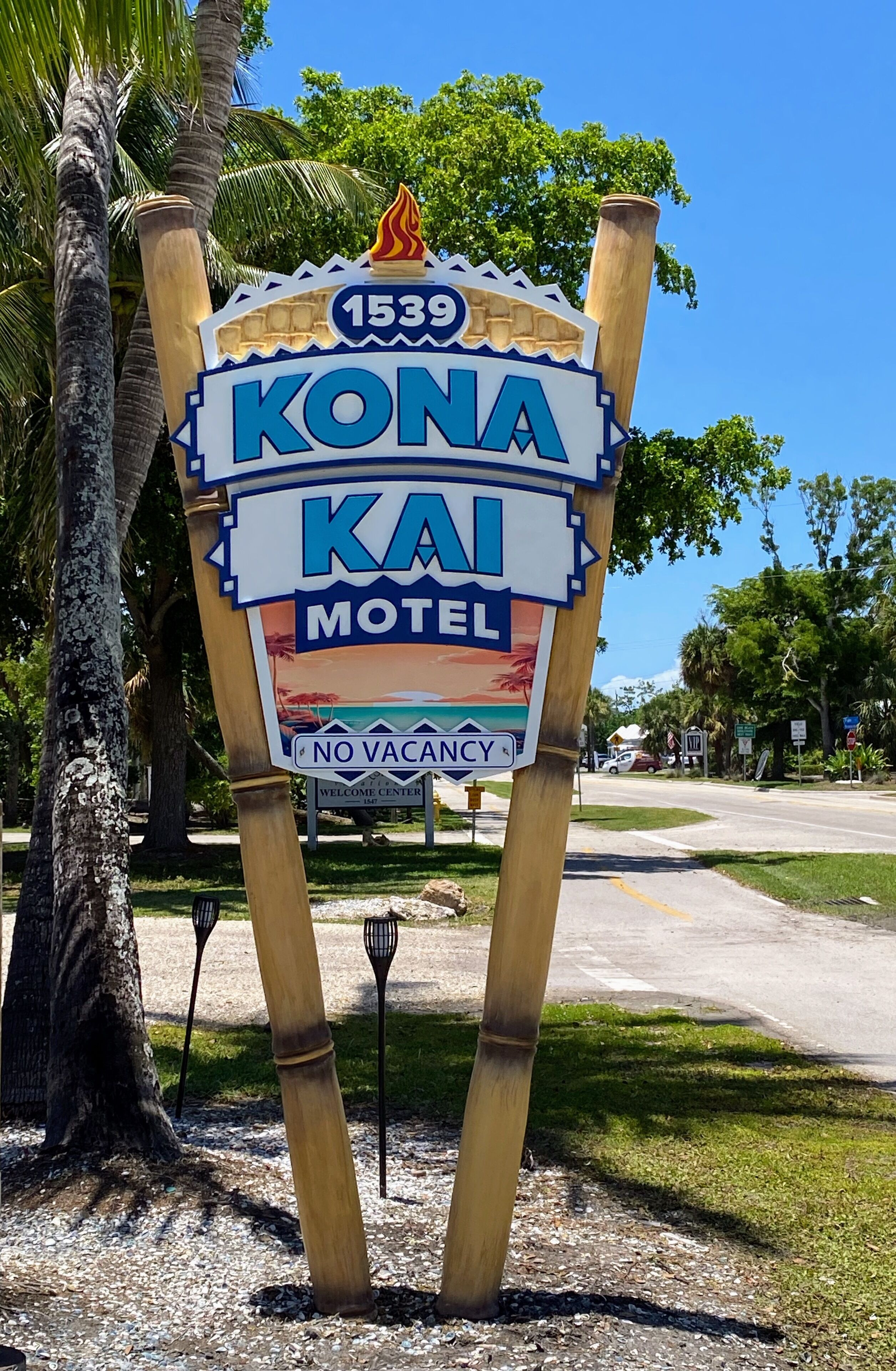 A sign for Kona Kai Motel with a 'No Vacancy' notice, located near a road with palm trees and a clear sky.