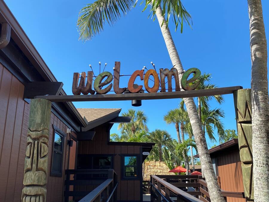 A wooden walkway leading to a building with a 'Welcome' sign, palm trees, and a thatched-roof structure in the background.