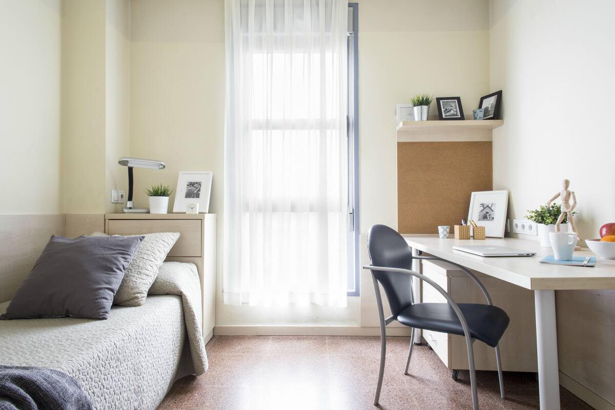 Een kleine, goed verlichte slaapkamer met een bed, een bureau en een stoel. Er is een raam met dunne gordijnen, een nachtkastje met een lamp, en een plank met decoratieve spullen.