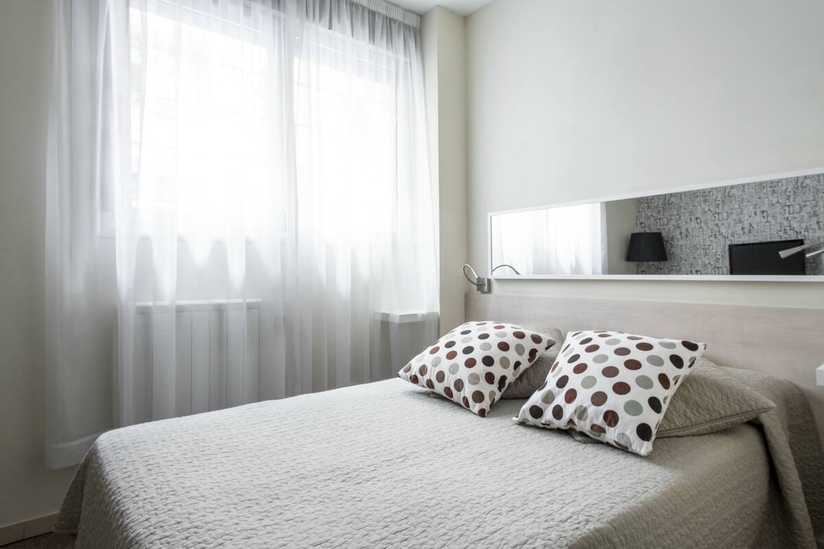 Een slaapkamer met een bed, twee kussens, een nachtkastje, een lamp en een raam met dunne gordijnen.