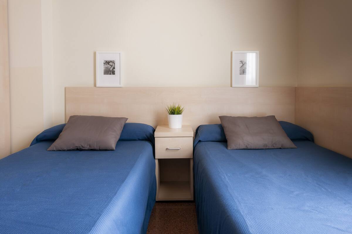 Een tweepersoonskamer met twee bedden, een nachtkastje, een kleine bloempot en twee ingelijste foto's aan de muur.
