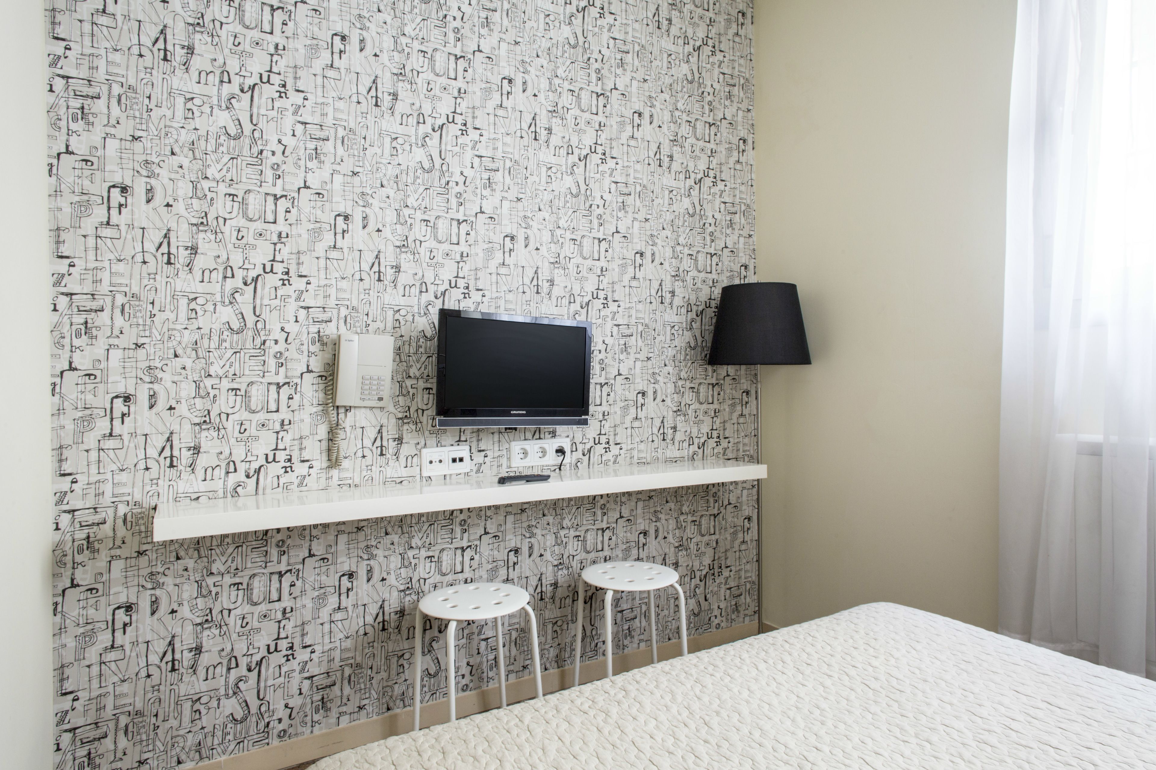 A hotel room with a patterned wall, a flat-screen TV, a black lamp, and two white stools.
