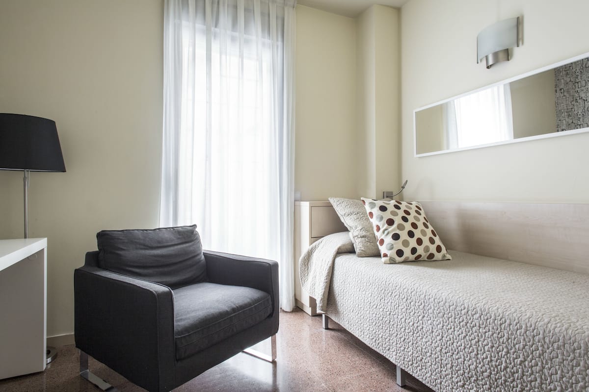 Een moderne slaapkamer met een grijze bank, een bed met gekleurde kussens, een nachtkastje en een grote spiegel aan de muur.