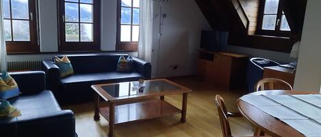 A cozy living room with a sofa, a coffee table, and a dining table. Large windows offer a view of the mountains.