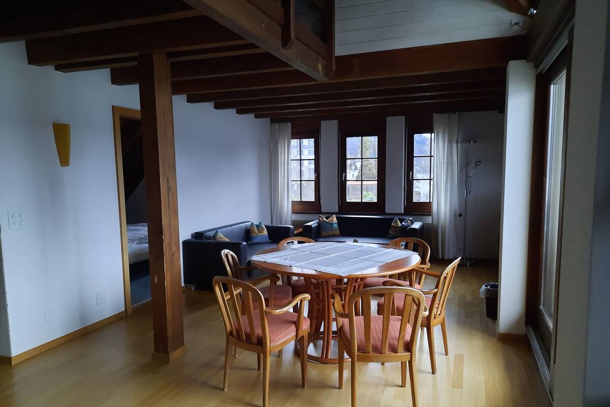 Un comedor con una mesa redonda de madera y sillas, un sofá con cojines y ventanas amplias con cortinas.