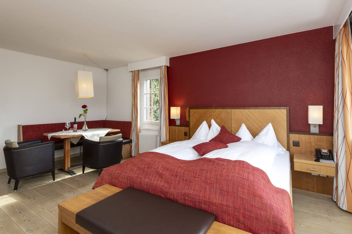 Una habitación de hotel con una cama grande, un escritorio, dos sillas, una mesita con un jarrón y una ventana con cortinas.