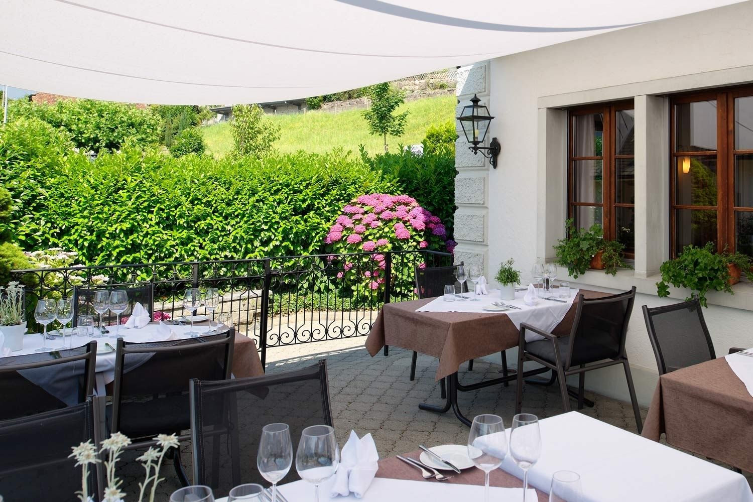 An outdoor dining area with tables set for two, surrounded by lush greenery and a pergola.