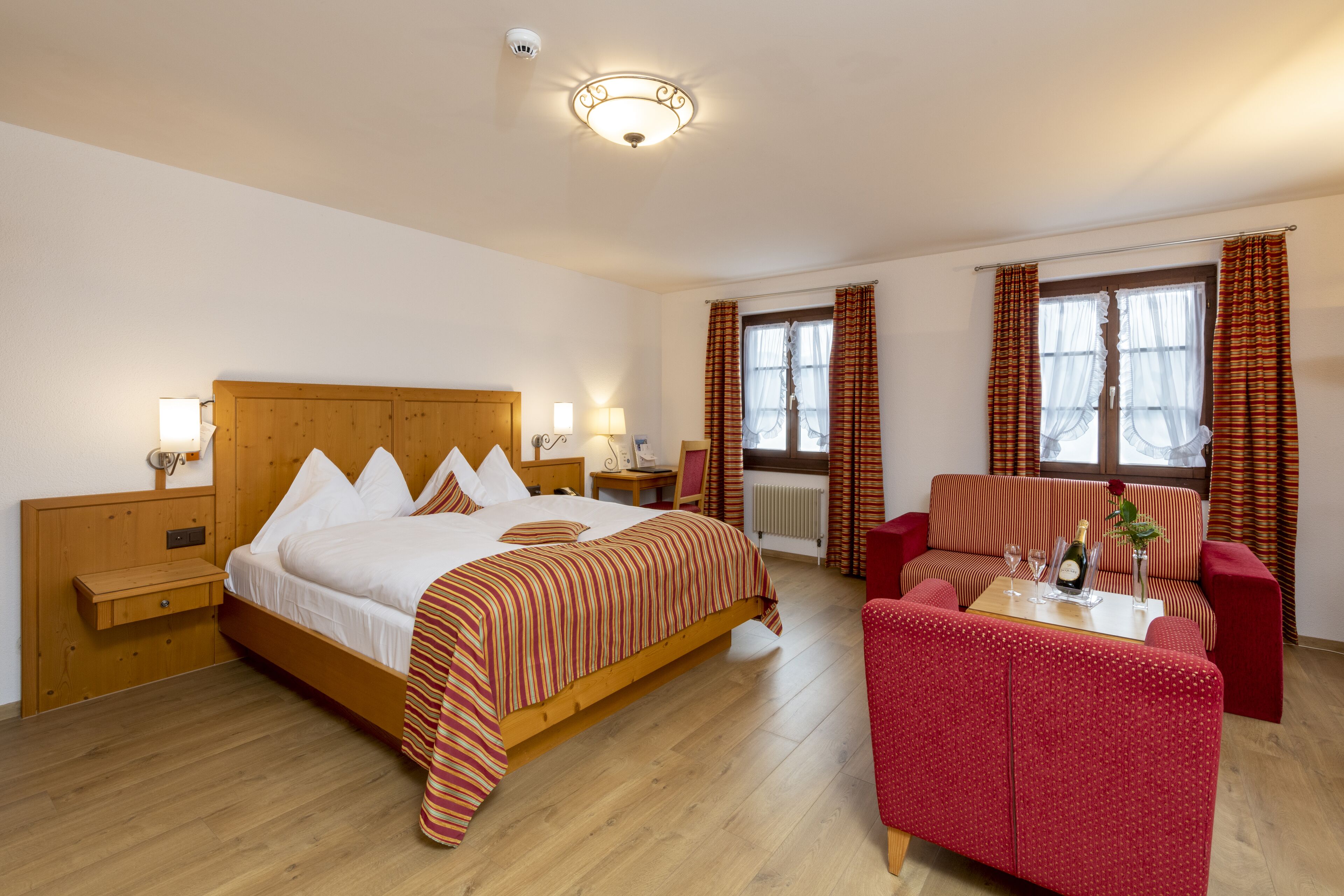 A hotel room with a wooden bed, a red armchair, a small table with a bottle and glasses, and a window with curtains.