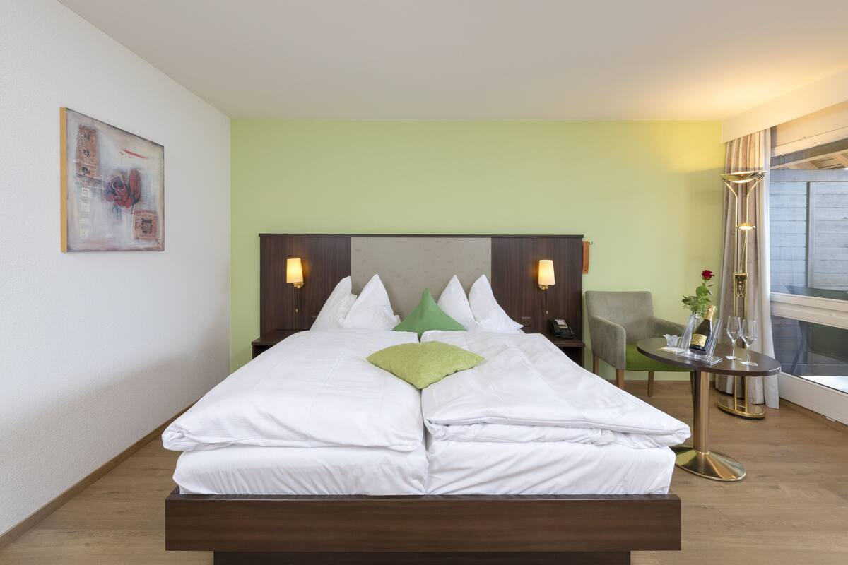 Una cama bien hecha con sábanas blancas y una almohada verde, una mesita de noche con una botella y vasos, y una silla en una habitación con paredes verdes y un ventanal grande.