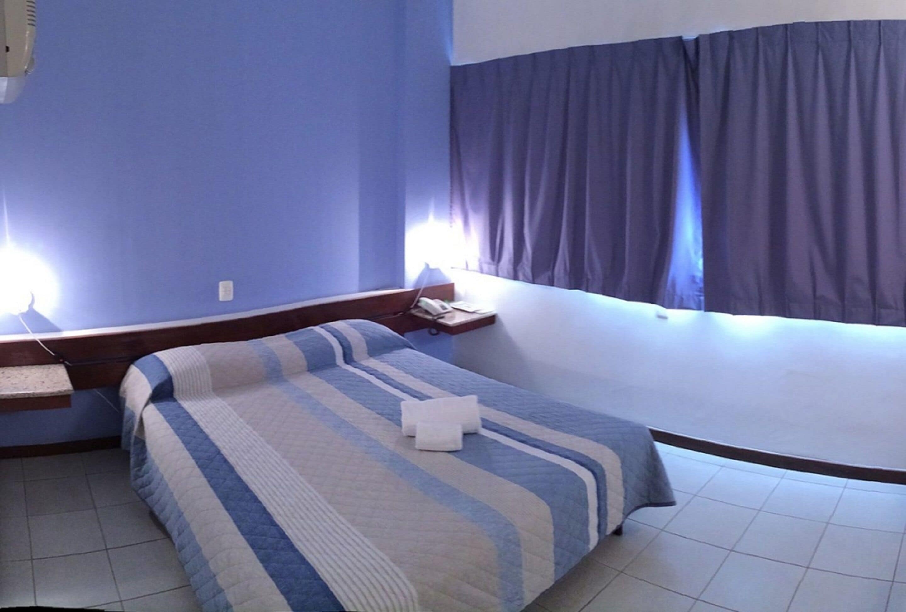 Une chambre d’hôtel avec un lit, une table de chevet, une commode, une lampe et une fenêtre avec des rideaux.