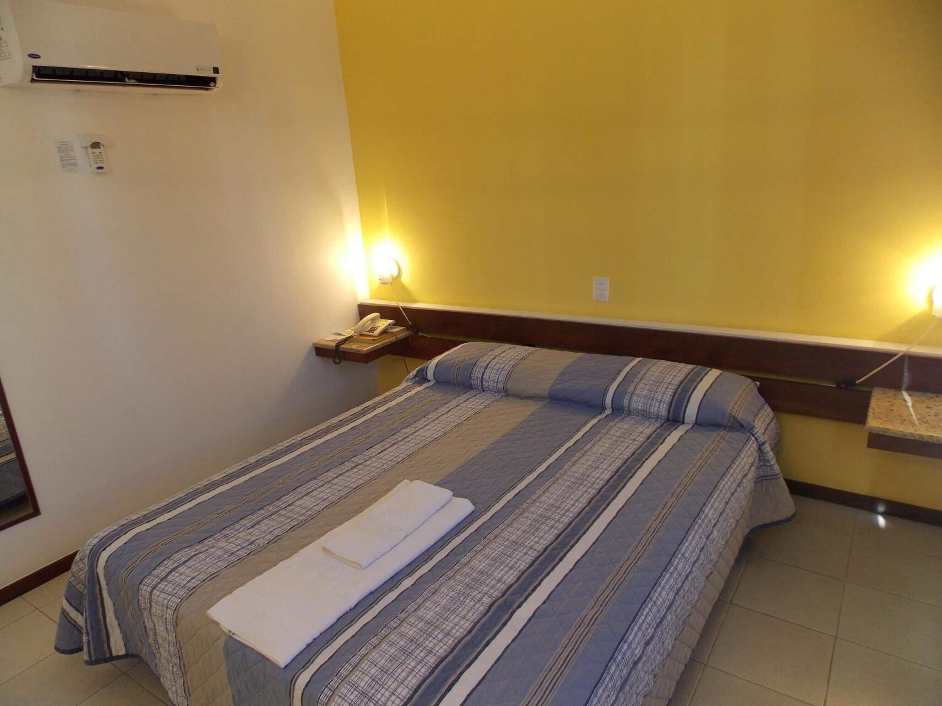Une chambre d’hôtel comprenant un lit, une table de chevet avec un téléphone, un climatiseur fixé au mur et un luminaire fixé au mur.