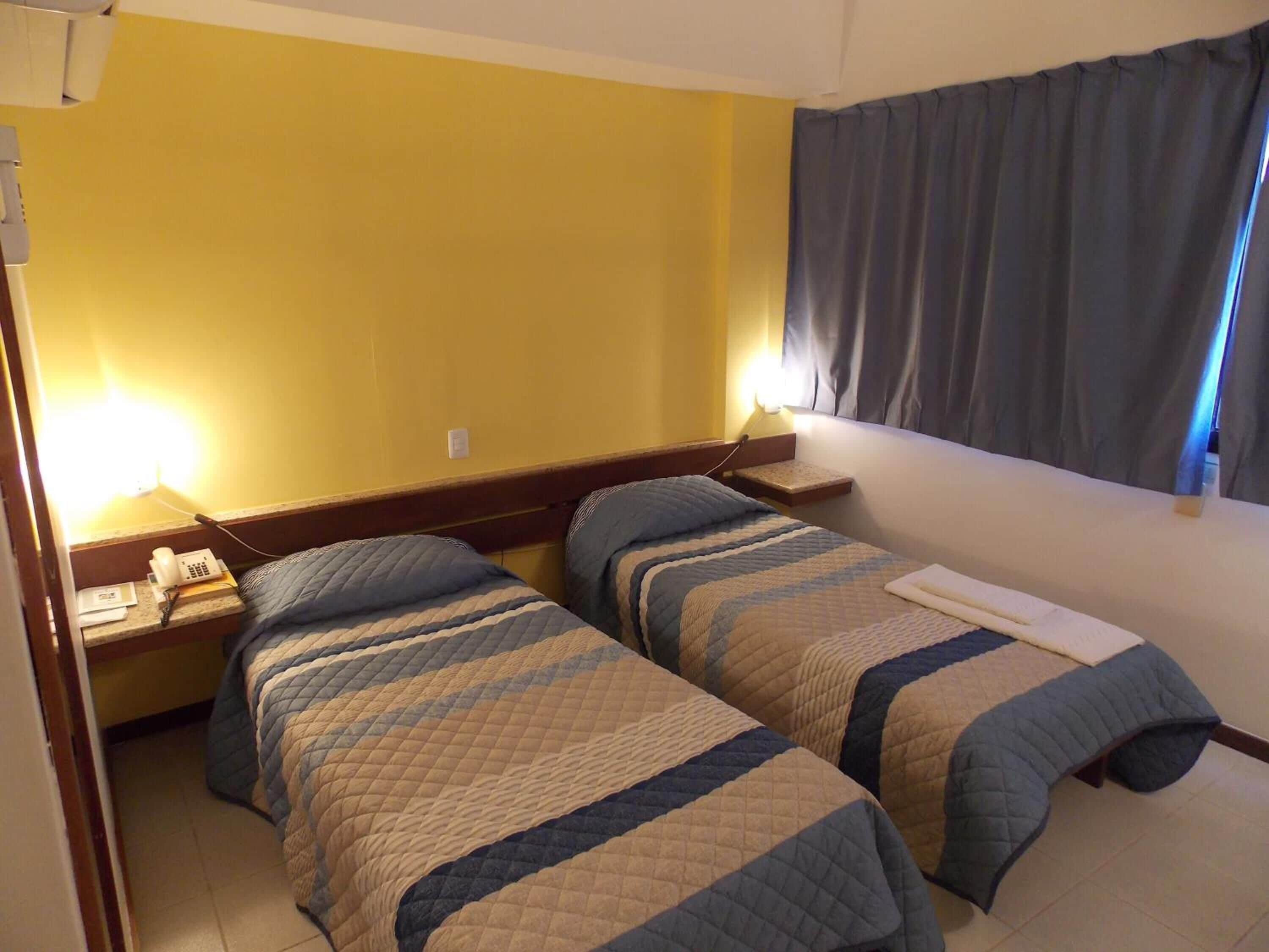 A hotel room with two beds, a bedside table with a phone, and a window with curtains.