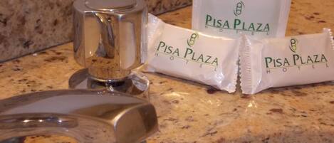 A bathroom sink with a faucet, a glass container, and two PISA PLAZA HOTEL toiletries packets.