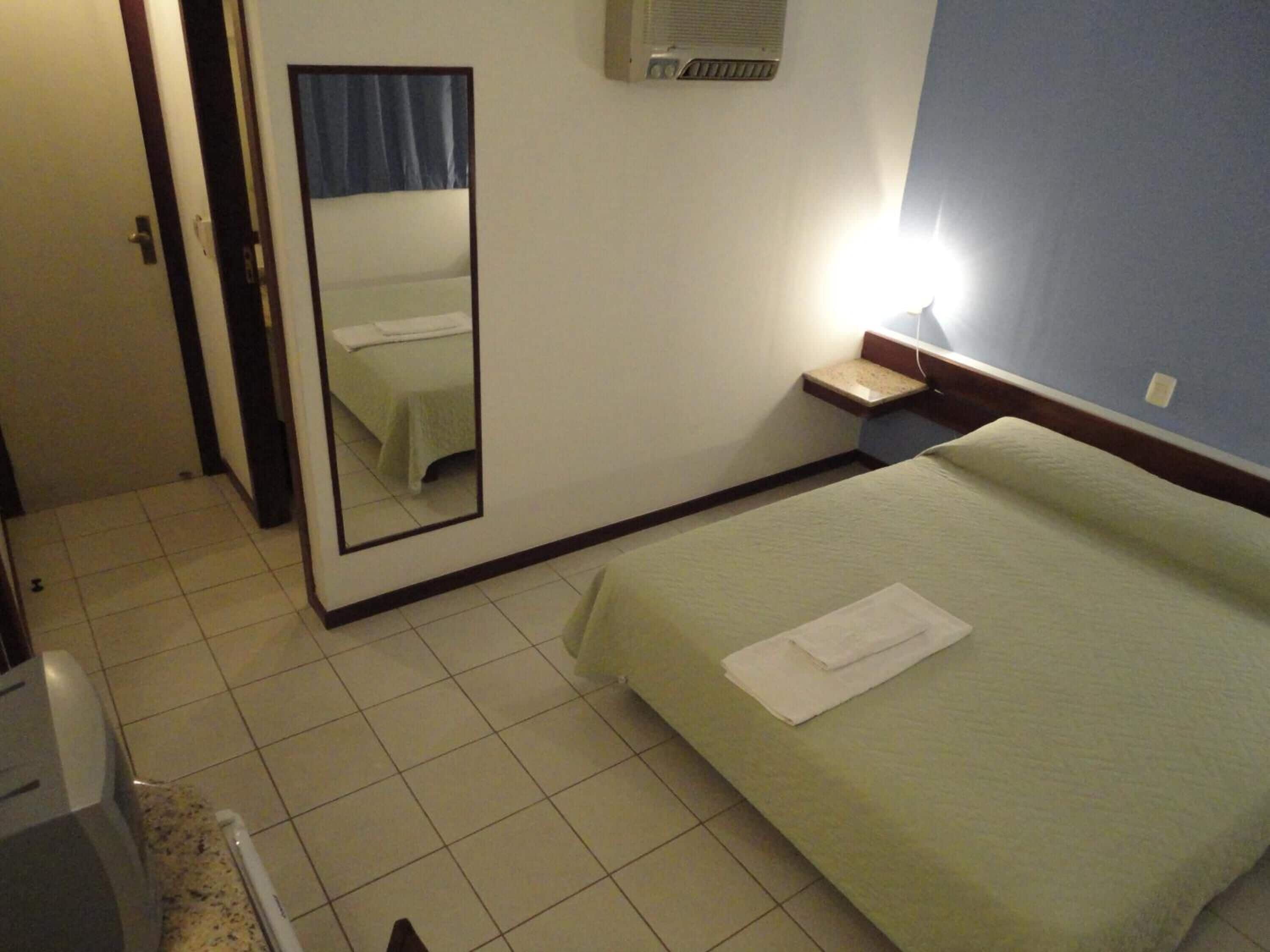Quarto de hotel com cama, espelho, mesa de cabeceira e uma pequena janela.