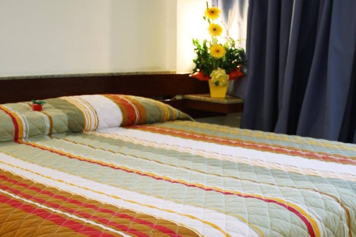 Una cama con un colcha multicolora, una mesita de noche con un florero y una ventana con cortinas oscuras.