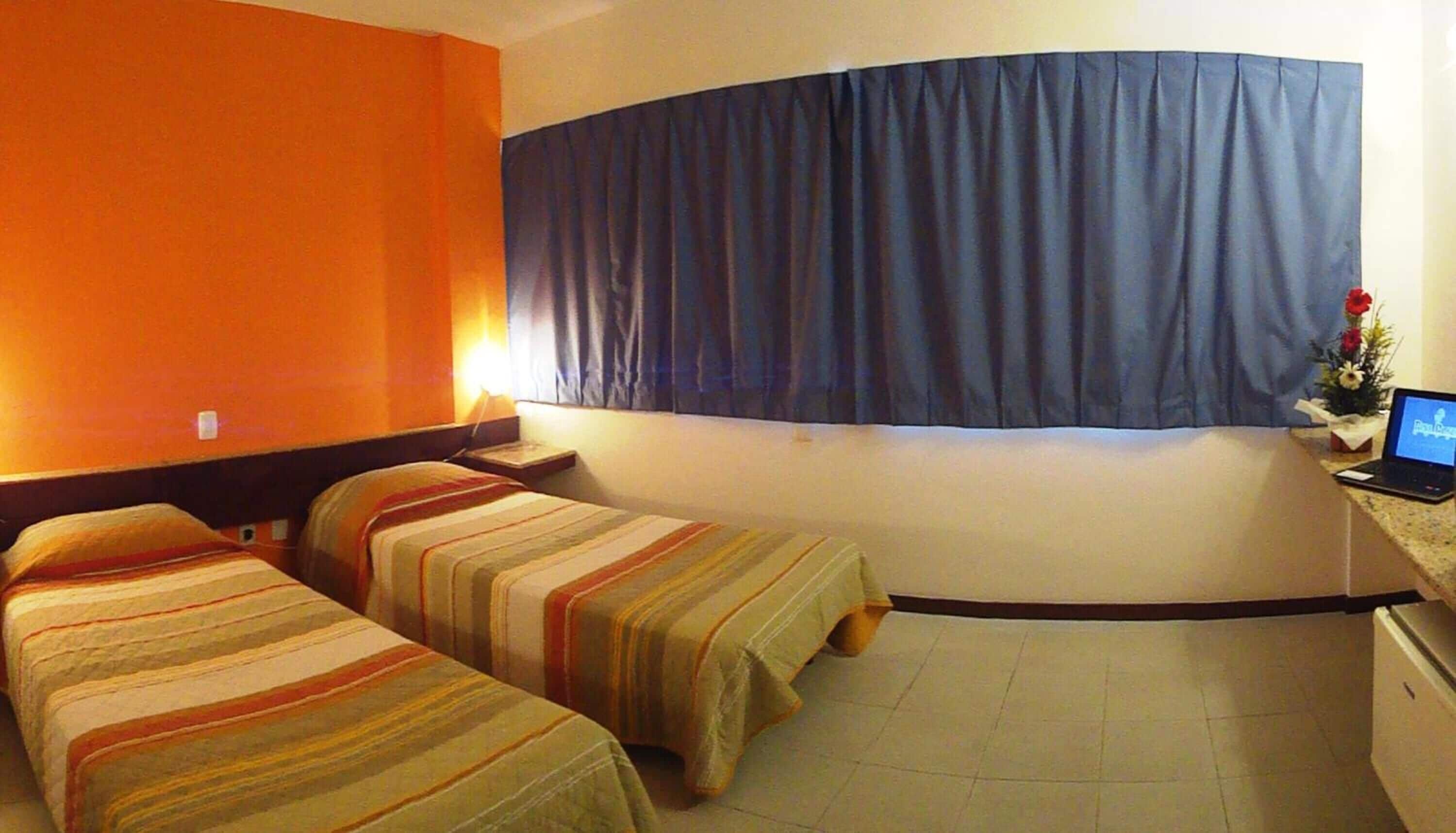 Habitación de hotel con dos camas, un escritorio con una laptop, una ventana con cortinas y una pequeña planta en el escritorio.